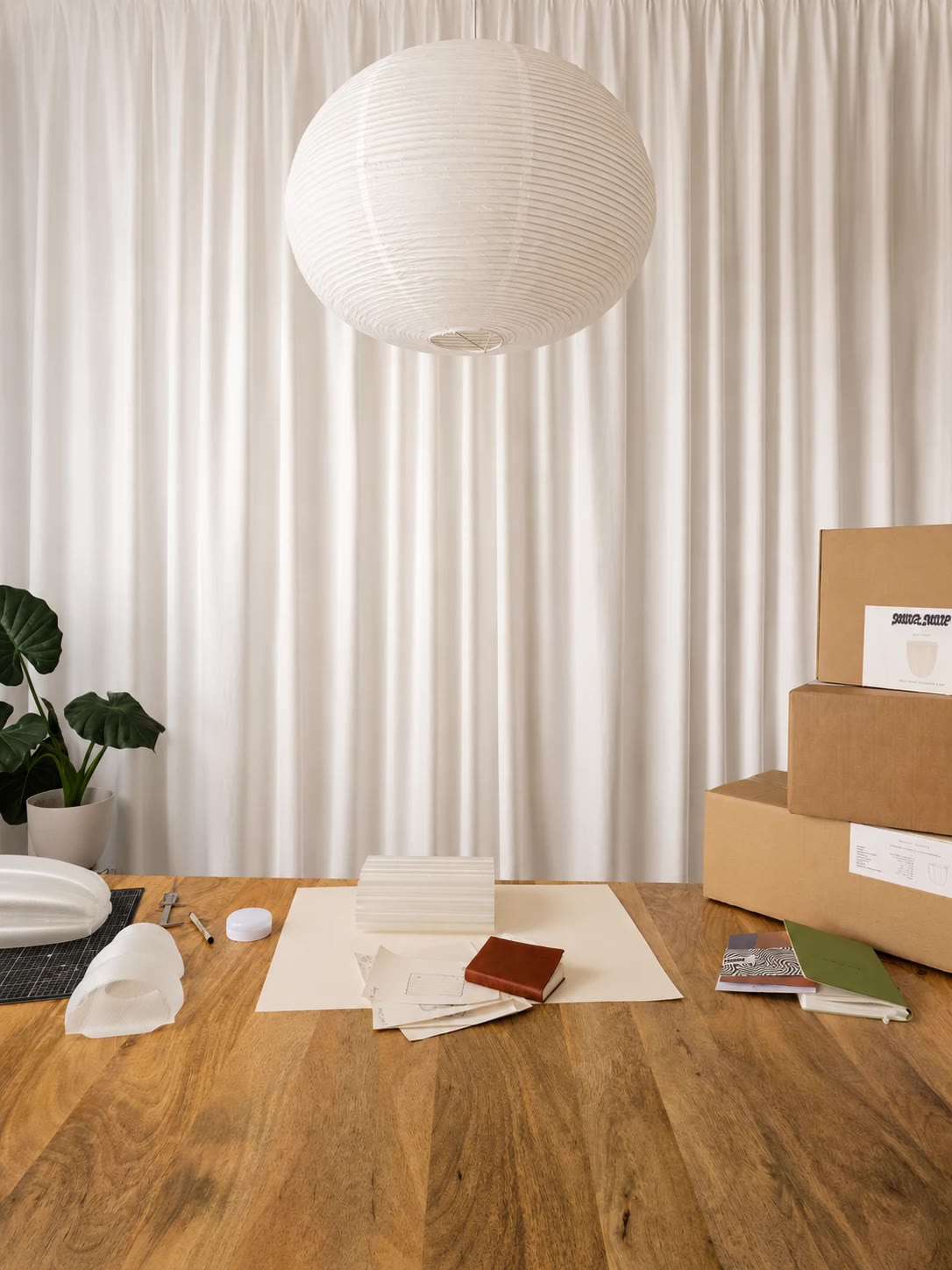 The studio table with lamp parts, sketches and packed Mur Mur boxes, under a paper lantern