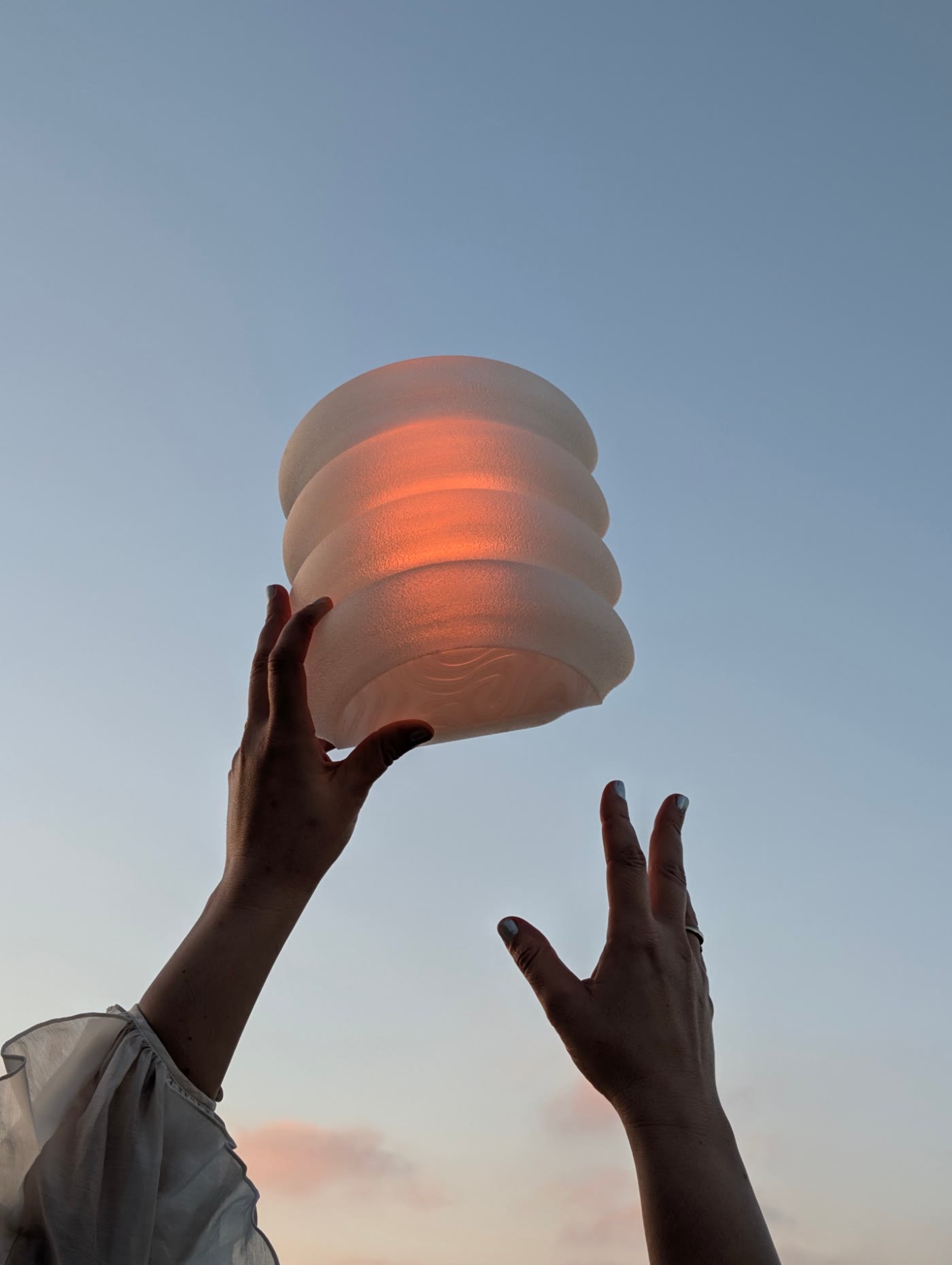 Two hands raise a glowing ribbed lamp against a sunset sky
